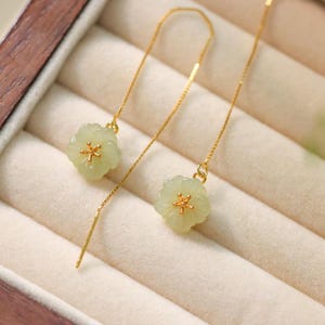 May include: Elegant gold earrings with pale green flower pendants and golden centres. The earrings have a long, slender chain design, presented in a jewellery box.
