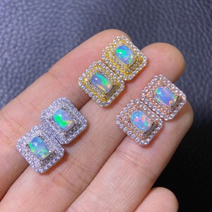 Puede incluir: Cuatro pares de pendientes de ópalo cuadrados. Cada pendiente presenta una piedra de ópalo central rodeada por un halo de pequeñas gemas transparentes. Los engastes son de oro, plata y oro rosa, mostrando los colores iridiscentes de los ópalos.