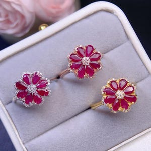 Può includere: Tre anelli a forma di fiore, con petali di rubino e dettagli di diamanti. Gli anelli hanno montature in argento, oro rosa e oro. Sono presentati in un portagioie grigio.