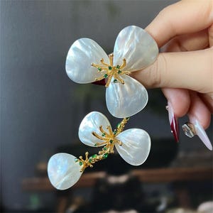 May include: A gold and white flower brooch with green gem accents. The brooch is made of mother of pearl and features a delicate floral design.
