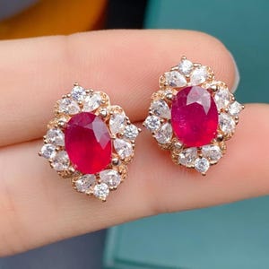 Puede incluir: Un par de pendientes de oro con una piedra preciosa de rubí rojo rodeada de piedras blancas más pequeñas.