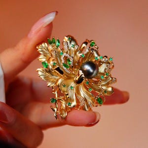 May include: A gold brooch in the shape of a flower with green emerald stones and a large black pearl in the centre.