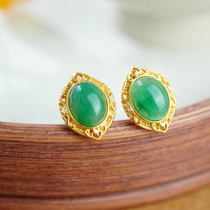 May include: A pair of gold-colored stud earrings. Each earring features a vibrant green oval-shaped stone set within a decorative gold frame. Small clear stones are set around the oval. The earrings are displayed on a wooden surface.