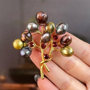 Op de afbeelding: Een broche van verguld metaal met een boomvormig ontwerp met een cluster van iriserende parels in grijstinten, bruin en goud. De parels zijn versierd met kleine rode edelstenen.