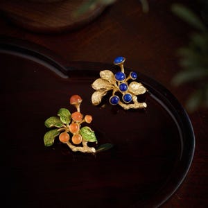Puede incluir: Dos broches dorados, uno con frutas naranjas y verdes, el otro con bayas azules. Los broches están detallados con hojas y tallos, sobre un fondo marrón oscuro.