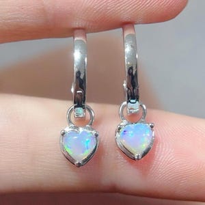 May include: A pair of silver hoop earrings with a dangling heart-shaped opal gemstone. The opal is iridescent with shades of blue, green, and pink.