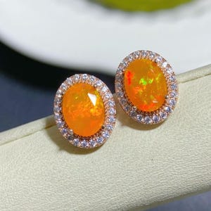 Può includere: Un paio di orecchini ovali con una grande pietra di opale arancione circondata da un alone di piccole pietre di zircone cubico trasparenti. Gli orecchini sono incastonati in un metallo color oro rosa.