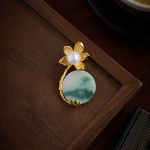 Peut inclure: Broche florale dorée avec une perle blanche au centre et une pierre de jade ronde verte et blanche. La broche comporte trois petites pierres vertes et est posée sur une surface en bois foncé.