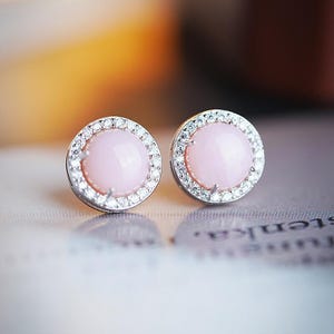 Puede incluir: Un par de pendientes de botón con un centro de piedra preciosa rosa claro y redondo, rodeado por un halo de pequeñas piedras claras y brillantes. Los pendientes tienen un engaste de metal plateado. Los pendientes se muestran sobre una superficie clara.
