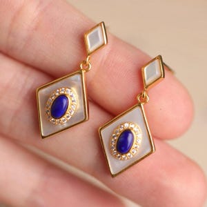 May include: A pair of gold earrings with a white mother of pearl diamond shape and a blue oval stone set in a gold frame with clear rhinestones.