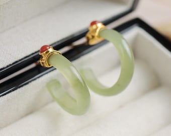 May include: A pair of hoop earrings featuring a light green jade design. The earrings have gold-colored accents and small red gemstones at the top. The earrings are displayed inside a black jewelry box with a white interior.
