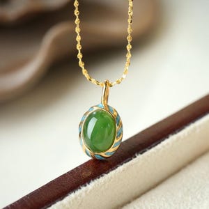 Op de afbeelding: Een gouden ketting met een groene jade hanger. De hanger is ovaalvormig en heeft een blauwe en gouden emaille rand.