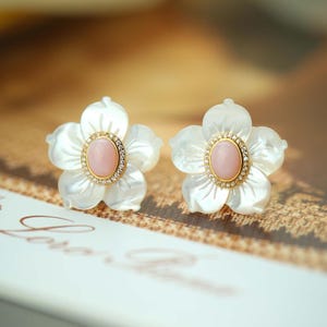 Può includere: Un paio di orecchini a forma di fiore bianco con pietre preziose rosa al centro e dettagli dorati. Gli orecchini sono su uno sfondo marrone chiaro.