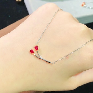 Op de afbeelding: Een delicate zilveren ketting met een gebogen staafontwerp, versierd met twee rode edelstenen. De ketting wordt op een hand gepresenteerd, wat de elegante en minimalistische esthetiek benadrukt. De rode edelstenen contrasteren.