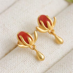 May include: A pair of gold earrings featuring red coral stones. The earrings are shaped like a flower with a teardrop dangling from the bottom.