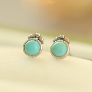 May include: A pair of silver-toned stud earrings. Each earring features a round, light turquoise stone set in a silver bezel. The earrings have a small, decorative element above the stone, adding a touch of detail.