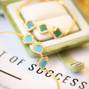 May include: Gold bracelets with clover-shaped pendants. One bracelet has turquoise-coloured stones, the other green. The pendants are outlined with small, clear stones. The bracelets are displayed on a white surface with the words "OF SUCCESS" visible.