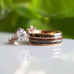 Könnte beinhalten: Ein Paar Eheringe. Der Verlobungsring hat einen großen, ovalen Diamanten, der in einer Roségoldfassung mit einem zarten Blattdesign sitzt. Der Ehering ist aus Roségold und hat eine schwarze und rote Einlage.