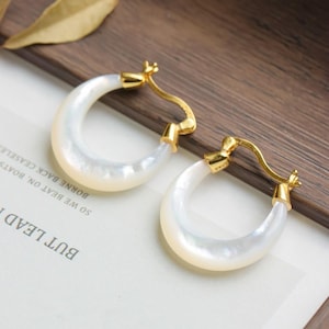 Peut inclure: Une paire de boucles d'oreilles créoles avec un fermoir doré et un corps blanc nacré en forme de croissant. Les boucles d'oreilles sont présentées sur une surface en bois avec un morceau de papier avec du texte.