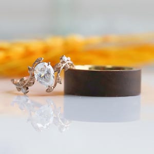 Peut inclure: Une délicate bague de fiançailles en or rose avec un diamant de forme poire serti dans un motif de feuilles. La bague est associée à une alliance en bois brun foncé.