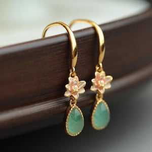 Peut inclure: Une paire de boucles d'oreilles dorées. Chaque boucle d'oreille présente une petite fleur rose et blanche au-dessus d'une pierre précieuse verte en forme de goutte. Les boucles d'oreilles sont présentées sur un fond marron foncé.