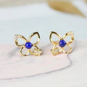 May include: A pair of gold-colored butterfly stud earrings. Each earring features a blue center stone, small clear stones, and a smaller butterfly design. The earrings are displayed on a light pink surface.