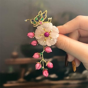 May include: A gold-toned floral brooch featuring a butterfly, a large flower, and pink bud accents. The butterfly has blue and green accents. The flower has a pink center and small clear stones. The pink buds hang from a gold stem.