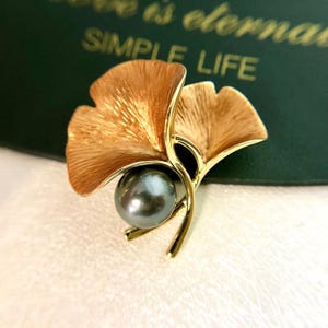 Op de afbeelding: Een goudkleurige metalen broche in de vorm van ginkgo bladeren, met een donkergrijze parel in het midden. De bladeren hebben een getextureerde afwerking. De broche staat tegen een groene achtergrond met de woorden "SIMPLE LIFE".