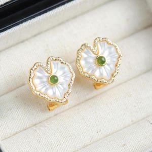 Puede incluir: Un par de pendientes de oro con un centro de concha blanca y piedra verde. Los pendientes tienen forma de flor.