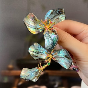 Pode incluir: Um par de grampos de cabelo em forma de flor, de cor verde e azul iridescente, com detalhes dourados e pedras preciosas vermelhas e verdes. Os grampos são feitos de concha de abalone e são projetados para parecer flores.