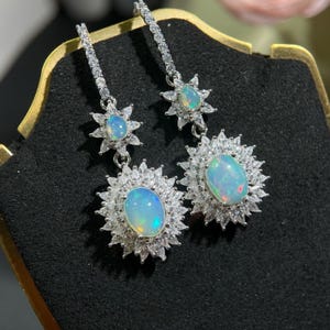 May include: A pair of silver dangle earrings with opal gemstones. Each earring features a large oval opal surrounded by a halo of smaller clear stones, and a smaller opal set in a star-shaped cluster of clear stones.
