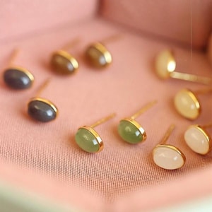 May include: A collection of oval-shaped stud earrings with gold-colored settings. The earrings feature various stone colors, including gray, green, and white. The earrings are displayed on a pink, textured surface.