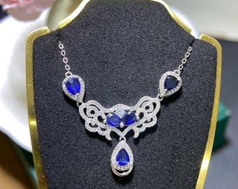 May include: A silver necklace with a series of blue gemstones and small clear stones. The necklace has a central design with multiple blue stones and a teardrop-shaped pendant. The necklace is displayed on a black jewelry stand.