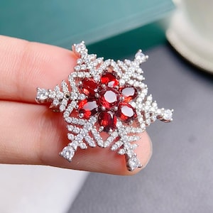 May include: A silver snowflake pendant featuring red gemstones. The snowflake is detailed with small white stones.
