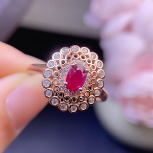 Può includere: Anello in oro rosa con un rubino ovale centrale, circondato da piccoli diamanti e un anello esterno decorativo. Il design mette in risalto il colore rosso del rubino e la brillantezza dei diamanti. Il design è intricato.