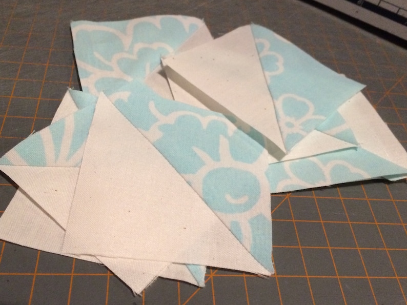 May include: A collection of white and light blue fabric squares, some with a floral pattern, arranged in a layered pattern on a cutting mat.
