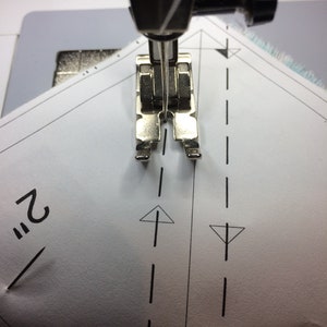 May include: A close-up of a sewing machine needle and foot positioned over a piece of white fabric with a dashed line and an arrow pointing upwards. The fabric is marked with the number "2" and a pin.