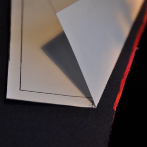 May include: A white piece of paper with a black border is folded over a piece of dark blue fabric with a red stripe. The paper is folded in a triangle shape.