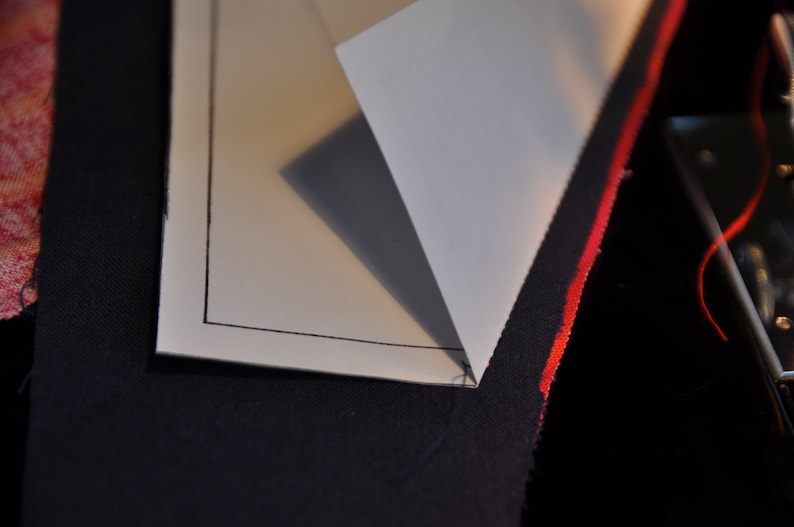 May include: A white piece of paper with a black border is folded over a piece of dark blue fabric with a red stripe. The paper is folded in a triangle shape.