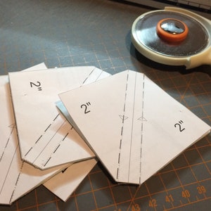 May include: Four white paper squares with 2" markings, stacked on a gray cutting mat with a rotary cutter in the background.