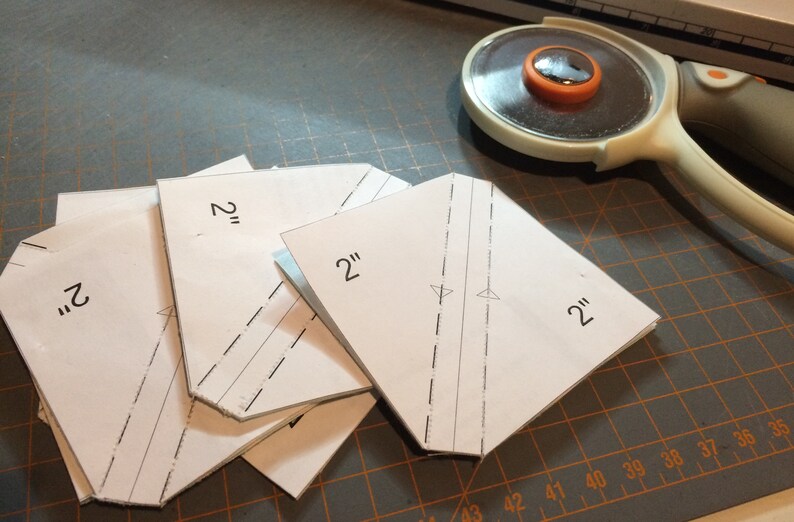 May include: Four white paper squares with 2" markings, stacked on a gray cutting mat with a rotary cutter in the background.