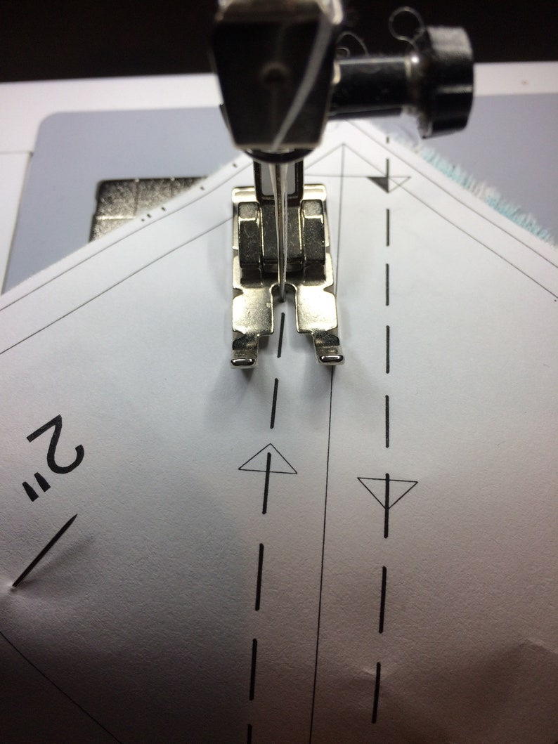 May include: A close-up of a sewing machine needle and foot positioned over a piece of white fabric with a dashed line and an arrow pointing upwards. The fabric is marked with the number "2" and a pin.