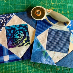 May include: Two blue and white patchwork quilt squares with a rotary cutter on a green cutting mat.