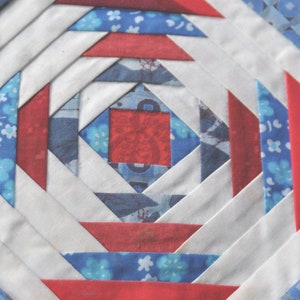 May include: A close-up of a red, white, and blue quilt block with a geometric pattern. The block is made of fabric squares sewn together in a pinwheel design.
