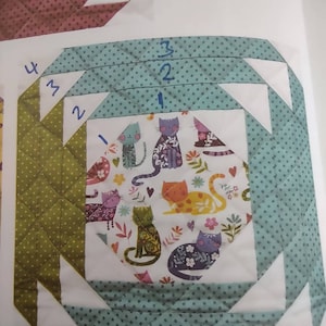 Pode incluir: Um bloco de colcha com fundo verde e branco e um centro branco com um padrão de gatos. O bloco é rodeado por uma borda turquesa com bolinhas brancas. Os números 1, 2, 3 e 4 estão escritos no bloco.