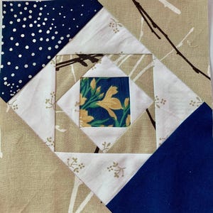 Puede incluir: Un bloque de edredón cuadrado que presenta un cuadrado central con un patrón floral amarillo sobre un fondo azul oscuro, rodeado de piezas de tela blancas y beige con motivos de ramas, y triángulos en las esquinas en azul marino con lunares y azul marino sólido.