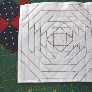 May include: A white fabric square with a brown grid pattern sewn on top. The grid pattern is made up of straight lines that form a series of concentric squares.