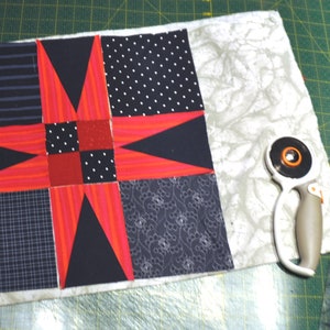 May include: A red and black patchwork quilt block with a star design. The block is on a white fabric background. A rotary cutter and scissors are on the right side of the image.