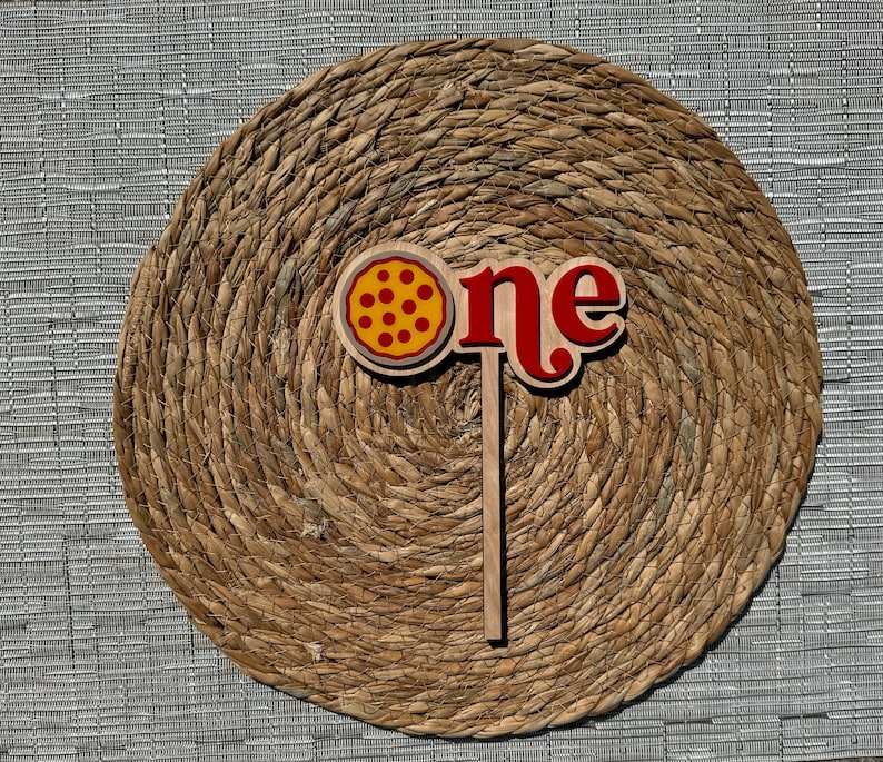 K&ouml;nnte beinhalten: Ein Holz-Kuchentopper mit dem Wort "one" in Rot und einem Pizzast&uuml;ck mit roten und gelben Pepperoni. Der Topper befindet sich auf einem braunen gewebten Hintergrund.
