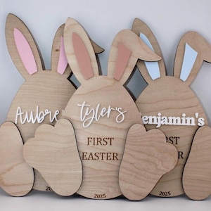 May include: Three wooden bunny-shaped signs with the names "Aubrey", "Tyler's", and "Benjamin's" and the text "First Easter" and "2025".